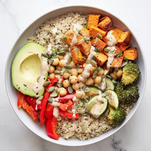 Colorful quinoa Buddha bowl with roasted sweet potato, chickpeas, and creamy tahini dressing on a white plate.