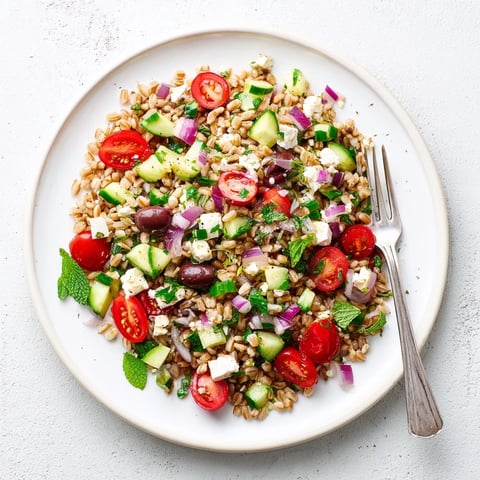 A close-up of the Farro Salad Mediterranean in a white bowl, showing chewy farro, vibrant red tomatoes, diced cucumbers, crumbled feta, and dark Kalamata olives on a rustic wooden table.  